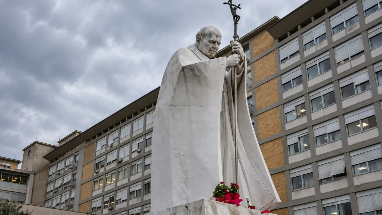 II. János Pál pápa kőszobra a római Gemelli Klinika épülete előtt, borús égbolt alatt. Illusztráció a XIV. Leó pápa tervezett tüdőműtétéről szóló hírhez.