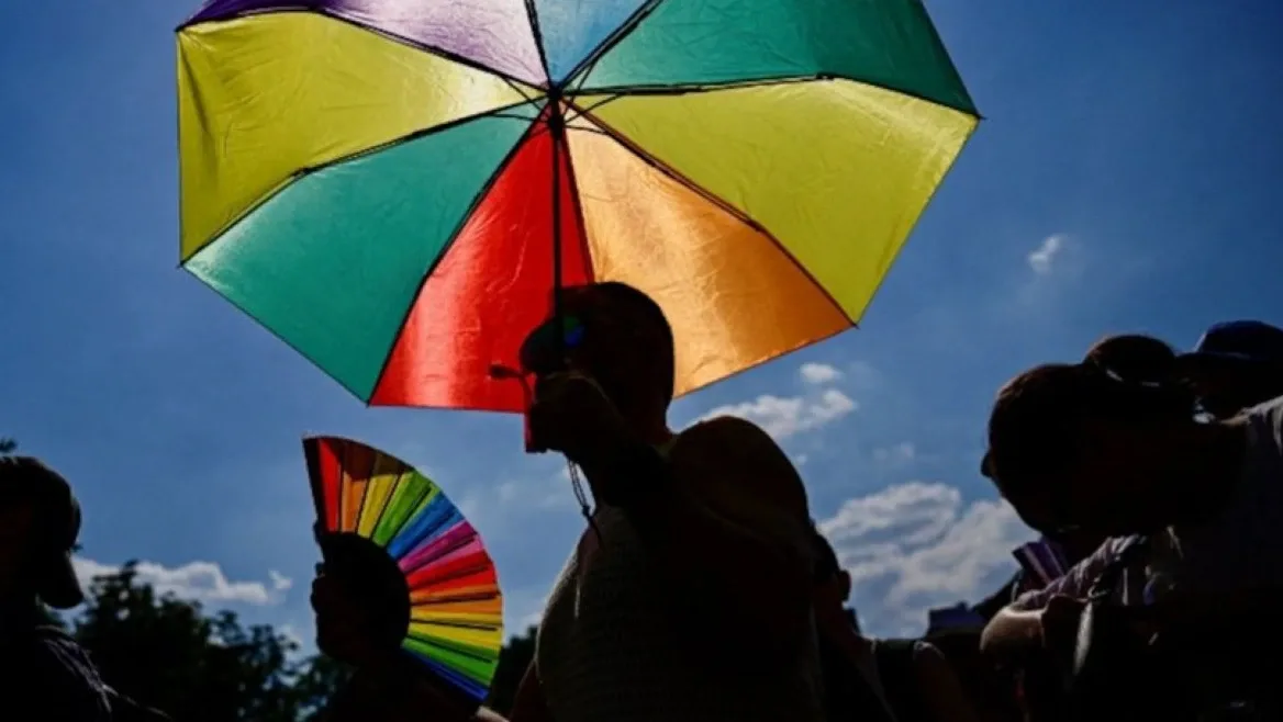 Szivárványszínű ernyőt és legyezőt tartó emberek egy szabadtéri rendezvényen, az LMBTQ-témájú szabályozás illusztrációjaként.