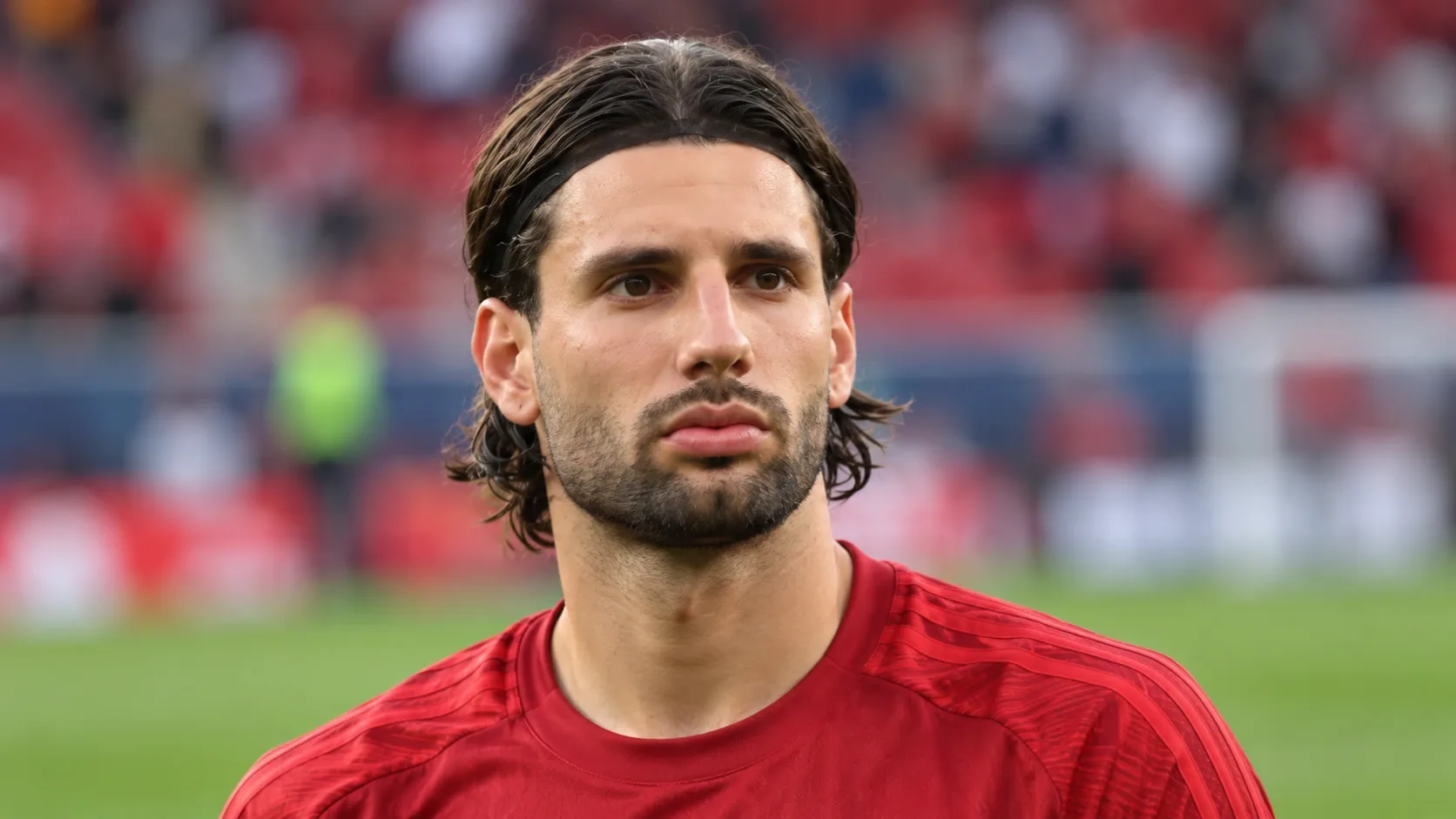 Szoboszlai Dominik piros sportmezben, elmosódott stadionháttér előtt. AI-generált portré.