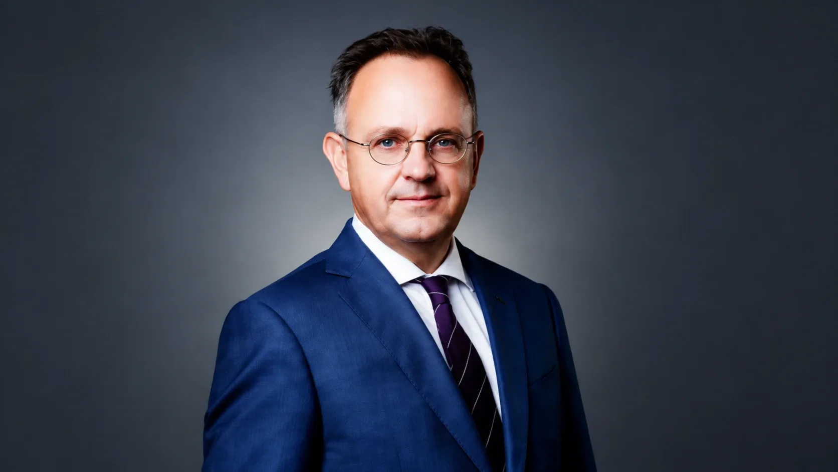 Bús Balázst ábrázoló portré kék öltönyben, fehér ingben és lila nyakkendőben, sötétszürke háttér előtt.