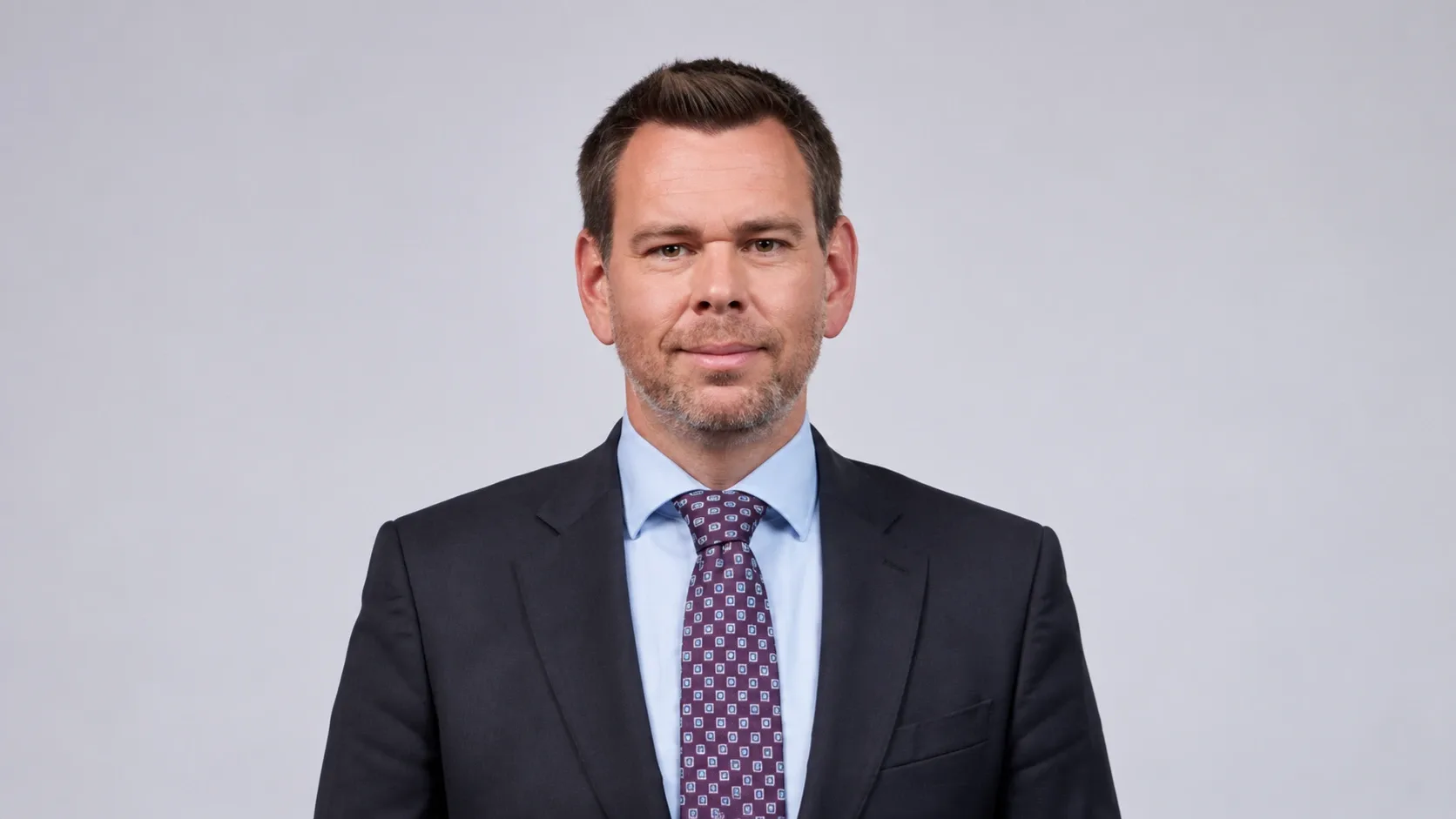 Vitézy Dávid sötét öltönyben, világoskék ingben és mintás nyakkendőben, világosszürke háttér előtt.