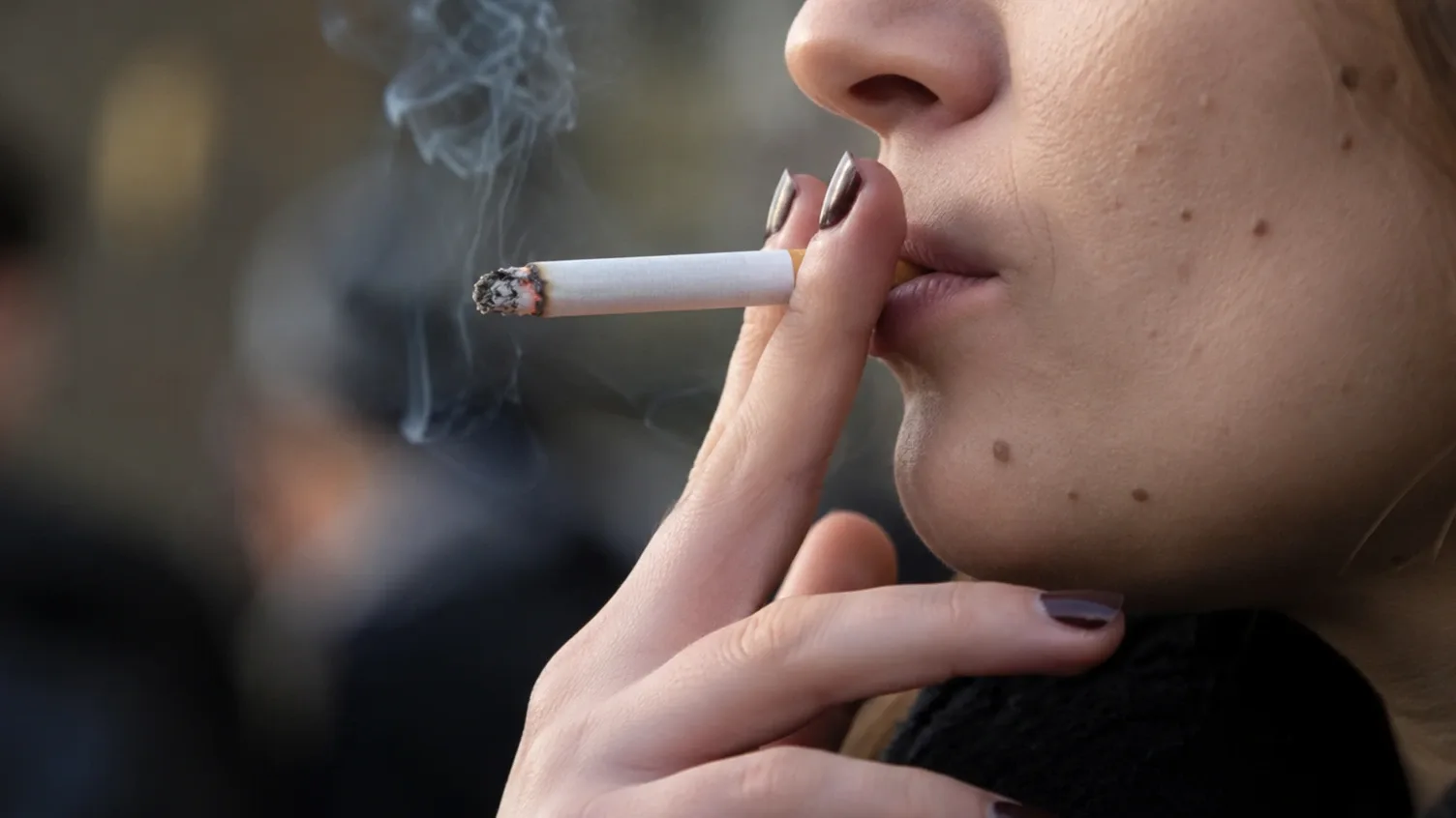 Közelkép egy égő cigarettát tartó és dohányzó nőről, a cigaretta drágulását és a dohánytermékek adóemelését illusztrálva.