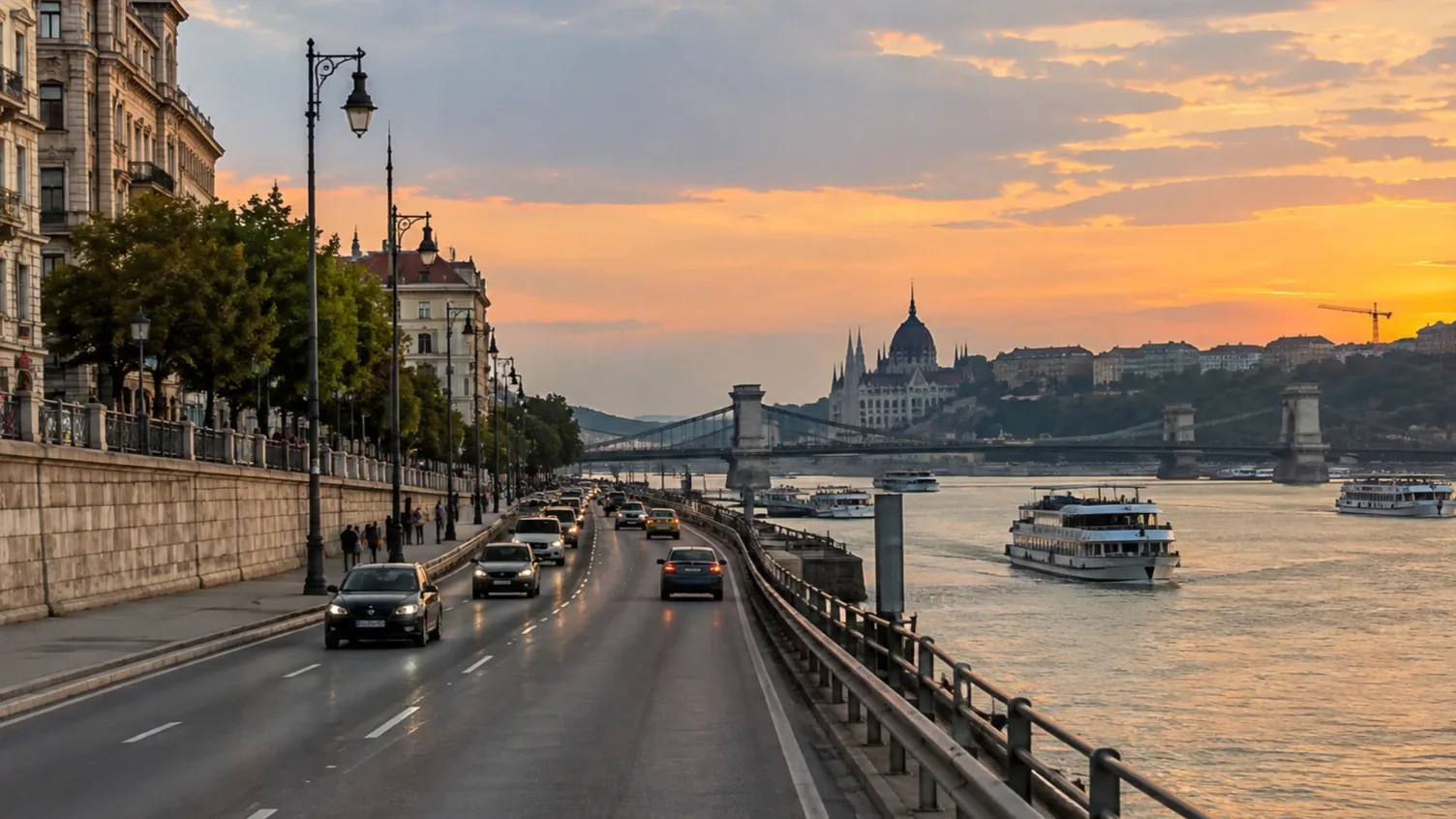 Autóforgalom a Pesti alsó rakparton a Duna-part mellett, háttérben Budapest ikonikus városképe naplementében.