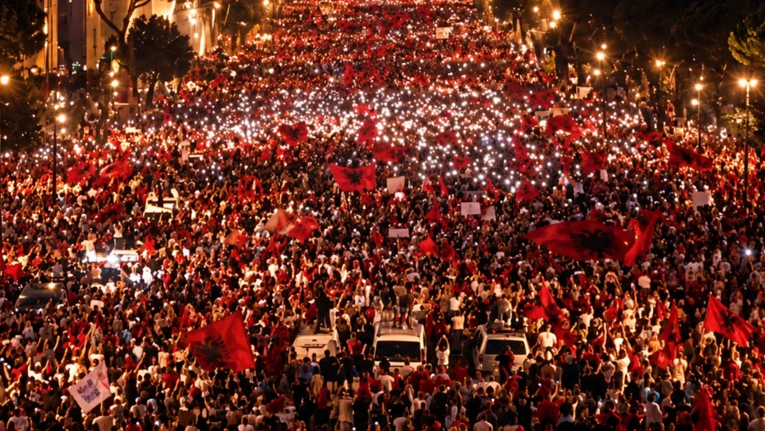 Hatalmas tüntető tömeg Tirana utcáin az esti órákban, albán zászlókkal és világító mobiltelefonokkal, a háttérben kivilágított városi épületekkel.