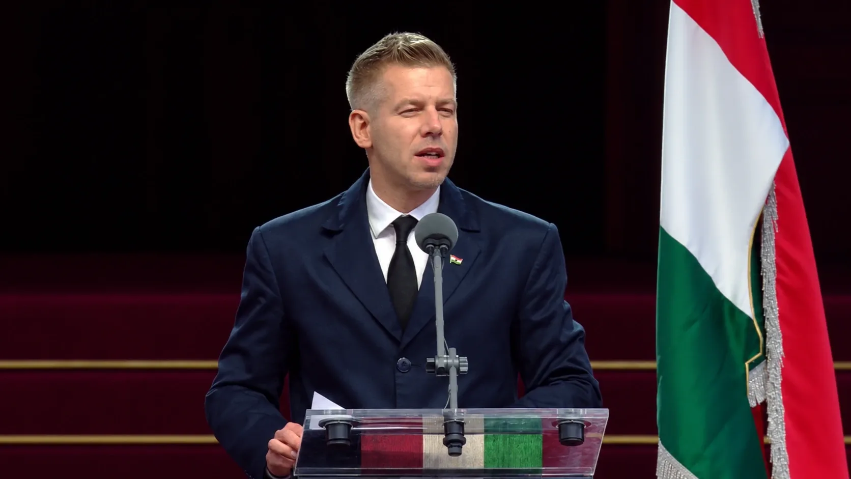 Magyar Péter sötétkék kabátban beszél a mikrofon előtt, mellette magyar zászló.