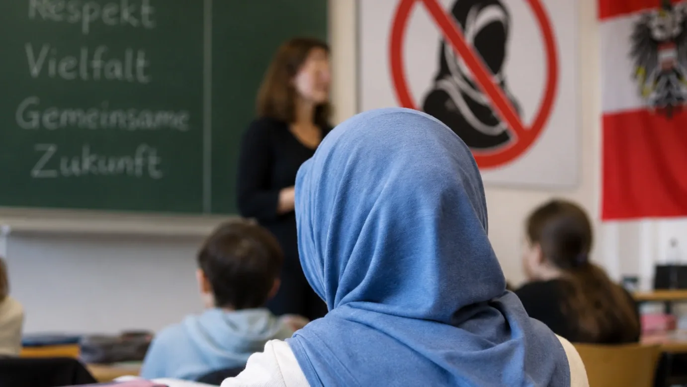 Muszlim diáklány kék fejkendőben egy osztrák tanteremben, a háttérben a hidzsábtilalomra utaló jelzéssel és az osztrák zászlóval.