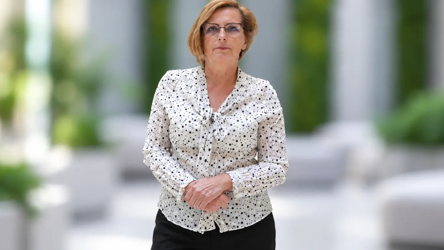 Dudás Katalin portréja világos, elmosott irodai háttér előtt, fehér pöttyös blúzban, összekulcsolt kézzel.