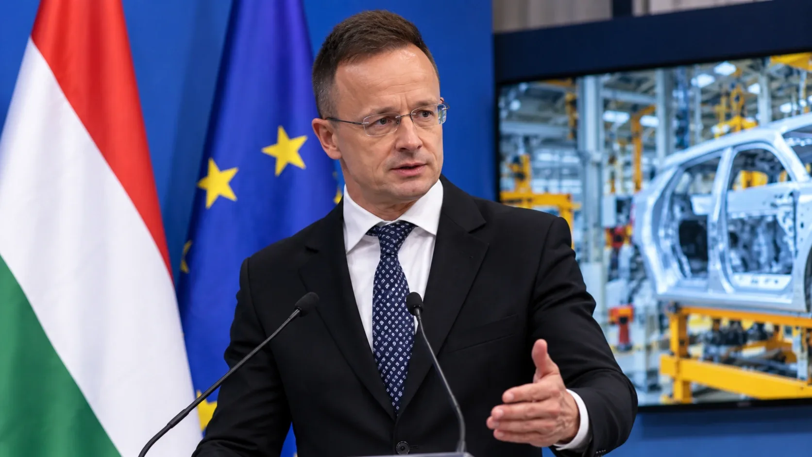 Szijjártó Péter egy autógyárat ábrázoló háttér előtt – illusztráció.