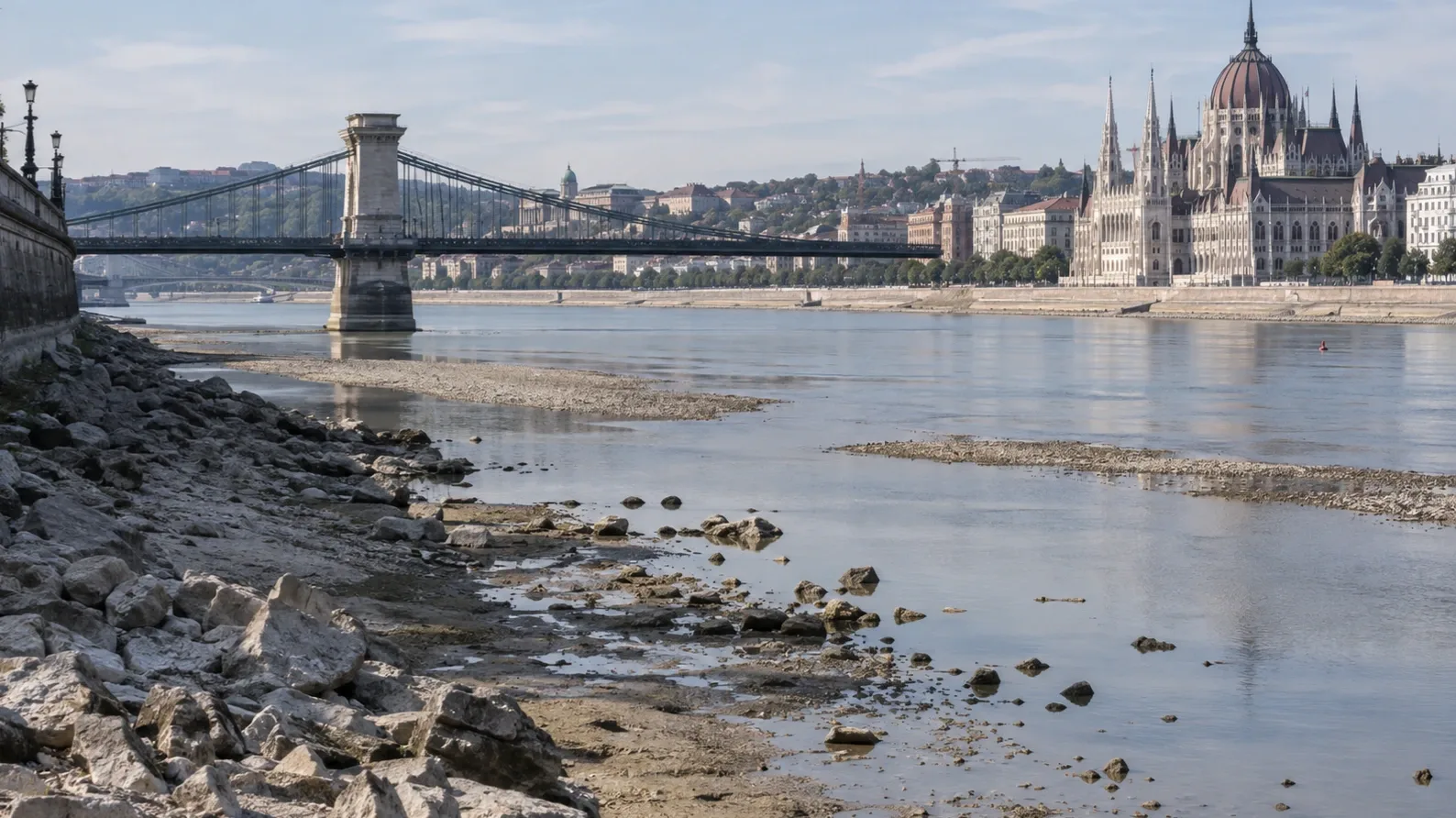 Rendkívül alacsony vízállású Duna Budapesten, előtérben a kiszáradt, köves parttal, háttérben a Lánchíddal és az Országházzal.